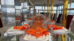 Una larga mesa de comedor dispuesta con varios vasos y servilletas naranjas, con vista a la ciudad a través de grandes ventanales.