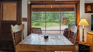 Ett rustikt matbord i trä med en klocka och ett ljus på, placerat framför ett stort fönster med utsikt över en skog.