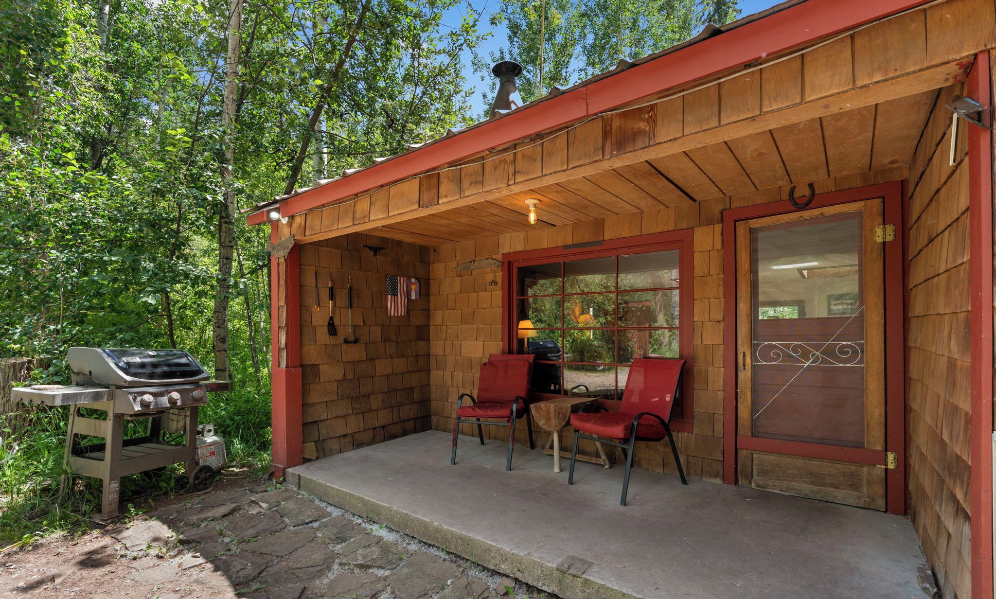 Une cabane en bois avec un porche, deux chaises rouges, un barbecue et un drapeau américain.