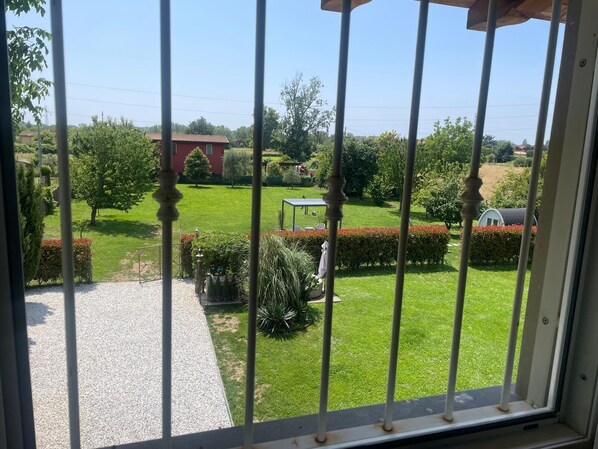 A view through a window with metal bars, featuring a well-maintained garden with a lawn, various shrubs, and a red building in the background. - Villa Francesca, pool (Capanne-Prato-Cinquale)