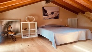 A cozy attic bedroom with a wooden ceiling, a bed with a blue bedspread, a radiator, a white shelf unit, and a wall-mounted picture of a rocky coastline. - Villa Francesca, pool (Capanne-Prato-Cinquale)