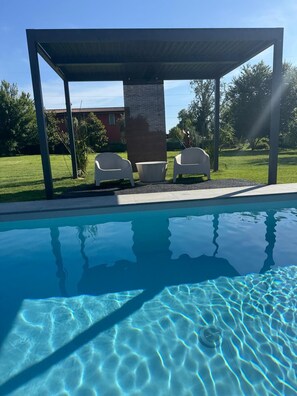 Une piscine à l’eau claire, entourée d’un patio abrité où se trouvent deux chaises blanches et un pilier en pierre.