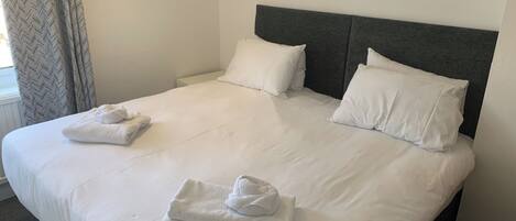A neatly made bed with white linens and pillows, a grey headboard, and a grey carpeted floor.