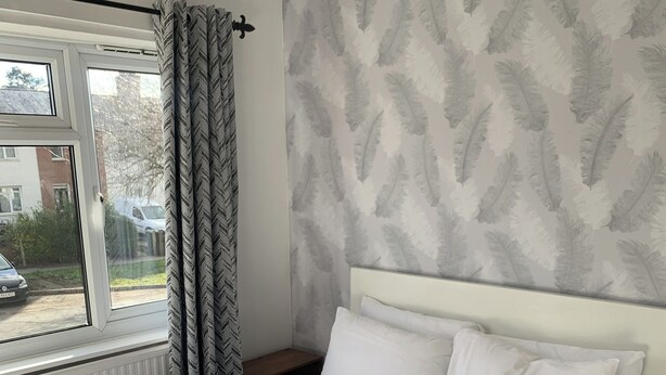 A neatly made bed with white linens, a window with curtains, and a patterned wall. - Ratcliffe House (Sileby)
