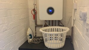 A compact laundry area with a front-loading washing machine, a white laundry basket, and a water heater with a digital display. - Ratcliffe House (Sileby)