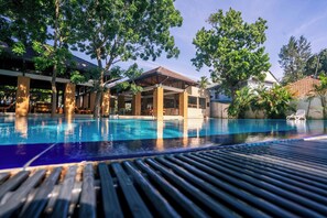 Ein Schwimmbecken mit Holzterrasse und Liegestühlen, umgeben von Bäumen und Gebäuden.