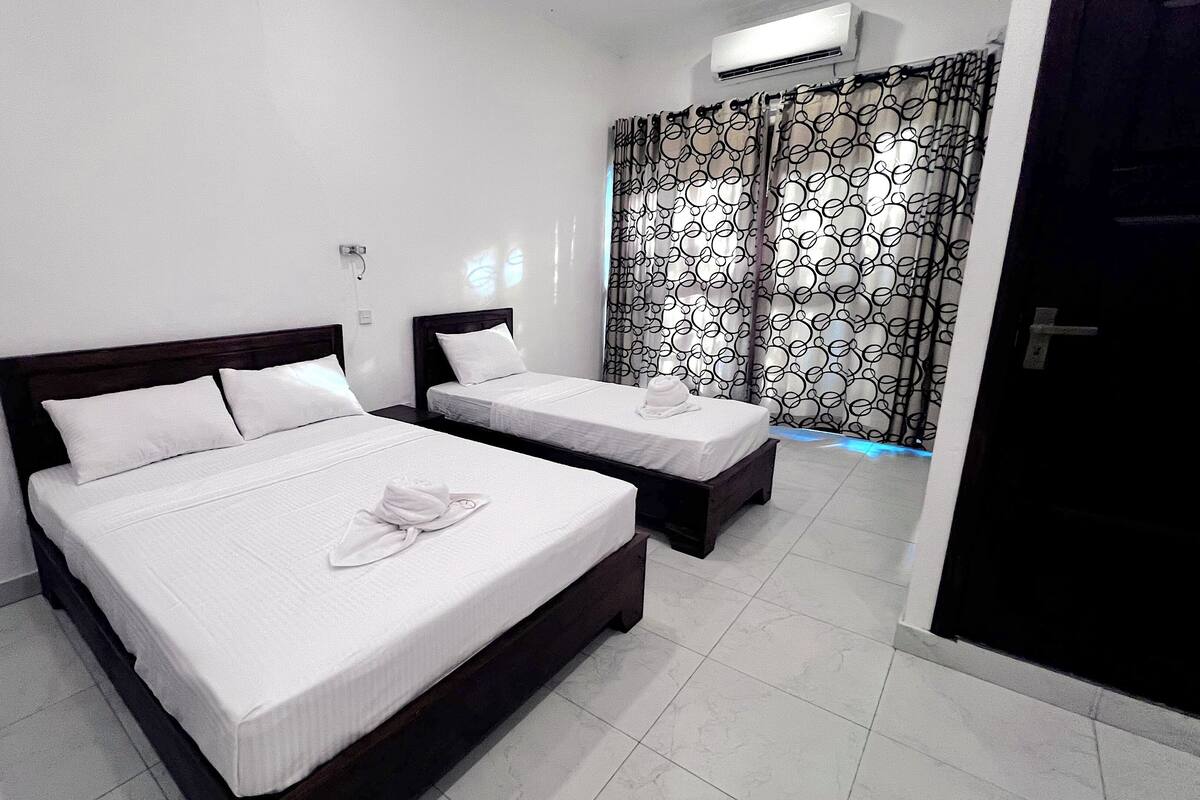 Ein Hotelzimmer mit zwei Betten, weißen Laken, einem hölzernen Kopfteil, Handtüchern auf den Betten und einem gemusterten Vorhang.