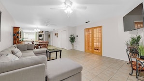A spacious living room with a grey sofa, a small table, and a ceiling fan. There is a wooden door and a TV mounted on the wall. - Cute, Comfy & Walkable 2/2 Condo Central McAllen (McAllen)