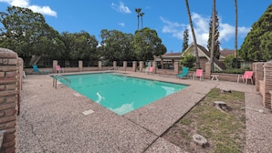 A rectangular pool with a clear blue water surface, surrounded by a pebble-covered deck. There are several lounge chairs, some with pink cushions, and tall palm trees in the background. - Cute, Comfy & Walkable 2/2 Condo Central McAllen (McAllen)