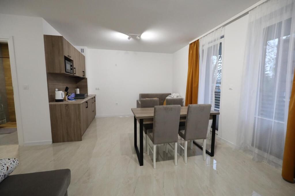 A modern living room with a dining area, a kitchen with wooden cabinets, and large windows with sheer curtains.