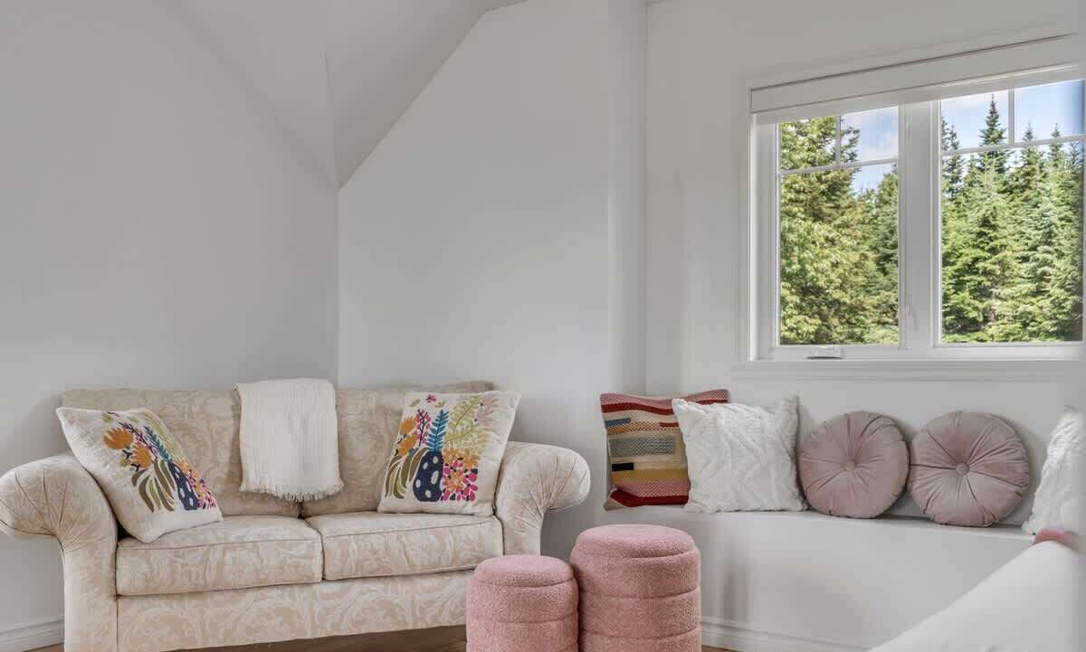 Una habitación con techo alto, una ventana que da a árboles, un sofá beige con cojines estampados y un sofá blanco con cojines a rayas.
