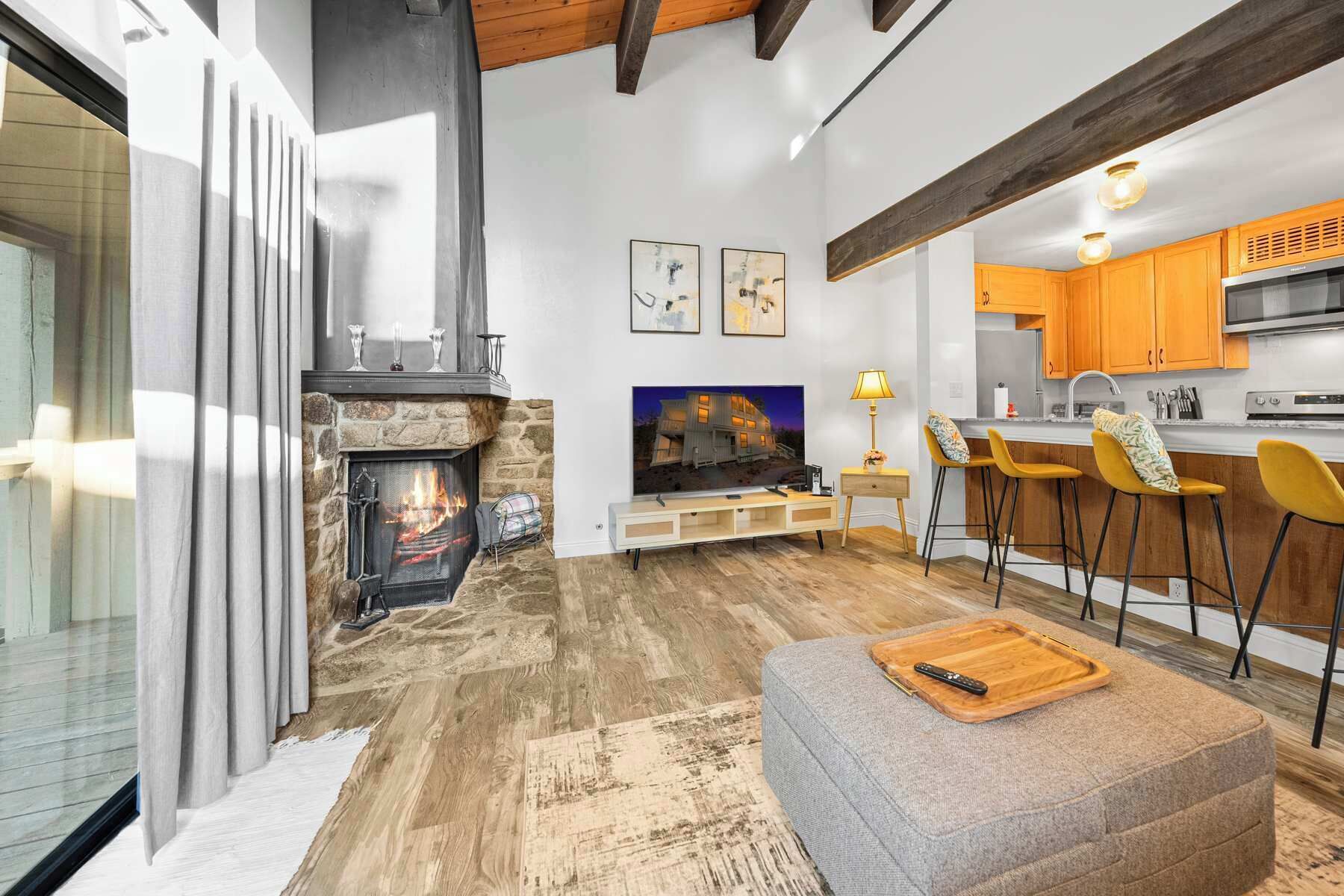 A modern living room with a stone fireplace, a TV, and a bar area with yellow chairs.