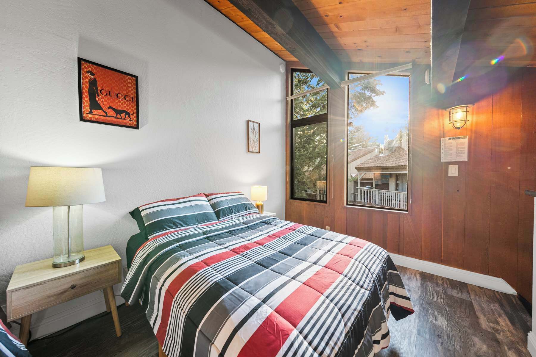 A bedroom with a large bed, two bedside lamps, a framed Gucci logo on the wall, and a window with a view of a building outside.