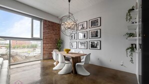 A dining area with a wooden table and white chairs, a chandelier, and black-and-white photographs on the wall. - Urban Opulence Loft | Rogers Place | UG Parking (Edmonton)