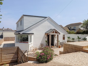 A single-story house with a sloped roof, a front porch with a bench, and a small garden area with pink flowers.