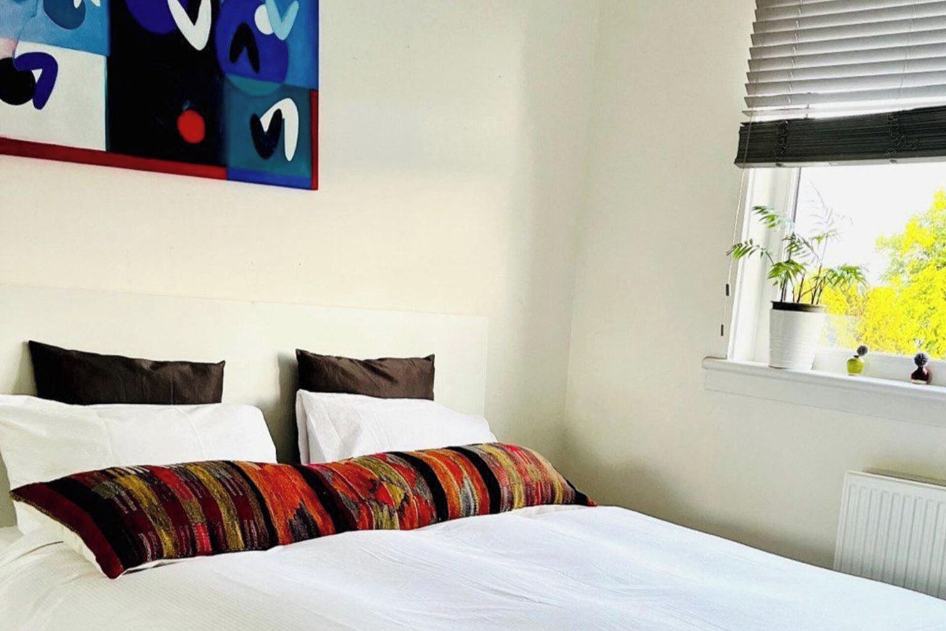 A bedroom with a bed, two pillows, a colorful blanket, a window with blinds, and a plant on the windowsill.