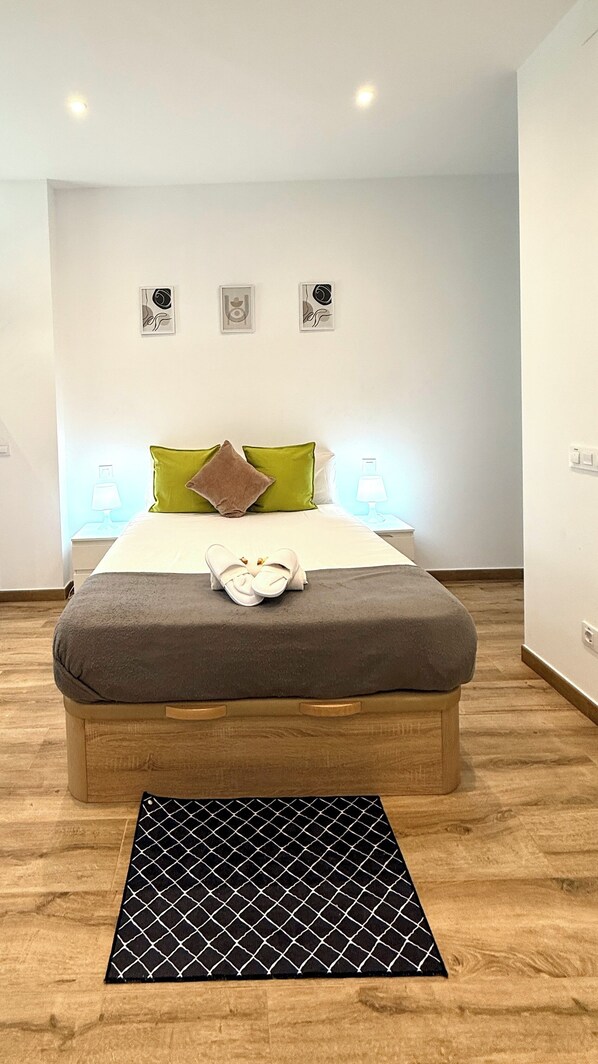 Un dormitorio ordenado con cama, almohadas, una mesita de noche con lámpara y una pequeña alfombra.