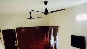 A single bed with a headboard, a bedside table, a chair, a ceiling fan, and a wall-mounted TV.