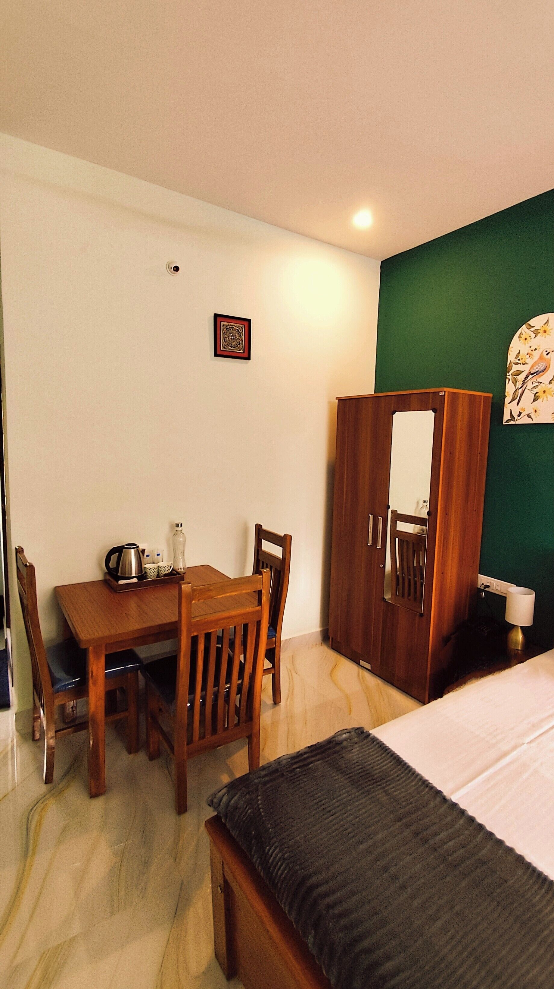 A room with a wooden dining table, chairs, a bed with a black headboard, a wardrobe, and a small framed picture on the wall.