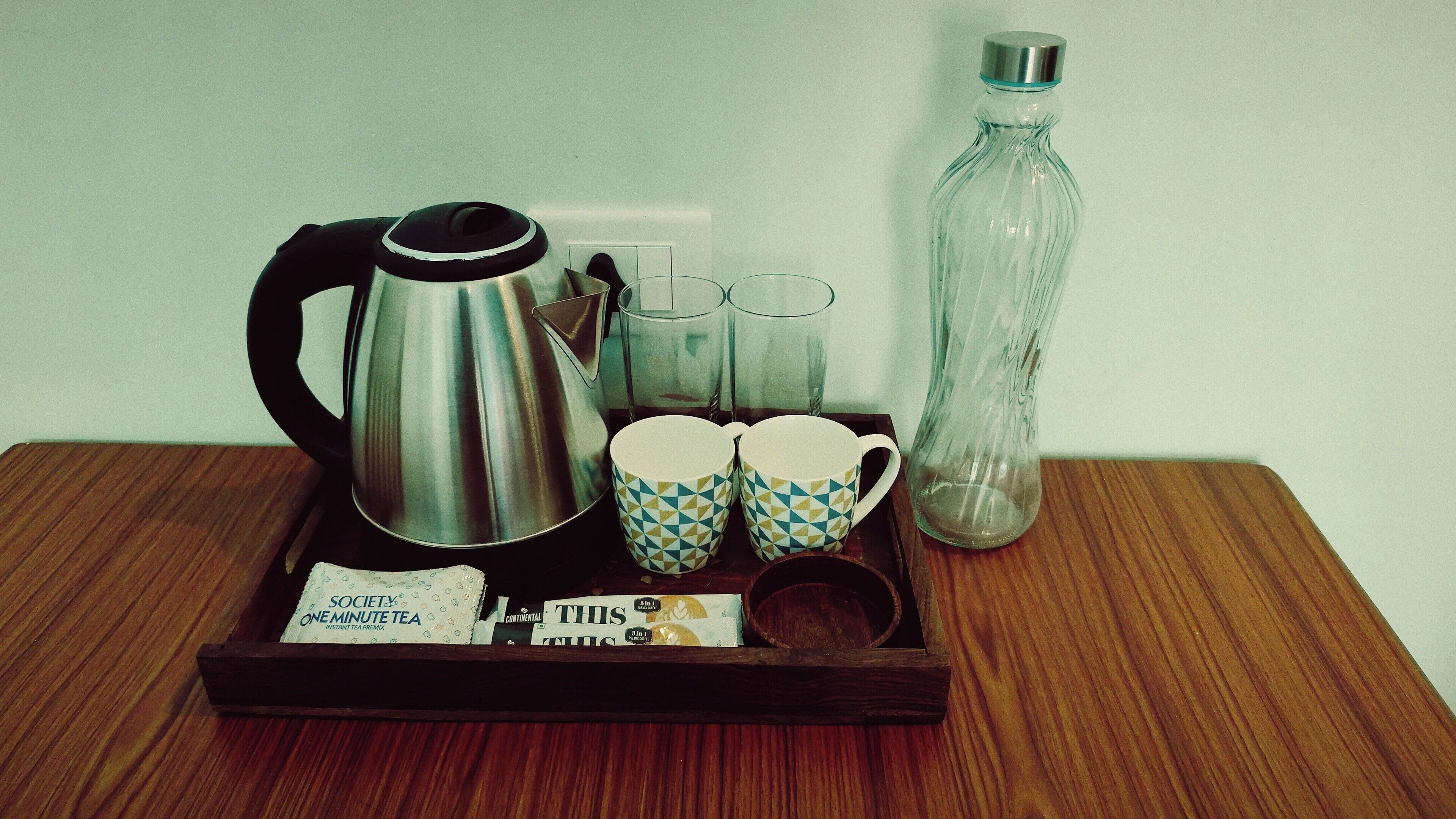 A tray with a stainless steel kettle, two patterned cups, a glass carafe, and tea packets on a wooden surface.