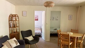 A cozy living room with a dark sofa, a wooden dining table, and a chair. There is a wicker shelf with decorative items and a bed with striped bedding in the background. - Evasion Nature Home & Glamping (Grayan-et-l'Hôpital)