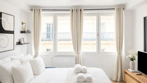 A hotel room with a bed, a TV, a window with curtains, and a view of the outside. - 1549 Anjou (Paris)