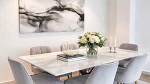 A dining area with a marble table, grey upholstered chairs with gold legs, a vase of flowers, and a large abstract painting on the wall. - 1549 Anjou (Paris)