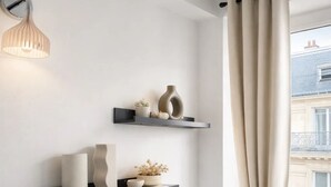 A minimalist room with two black floating shelves, each holding decorative items and a rolled-up towel. A modern pendant light hangs on the left wall, and a window with a curtain is visible on the right. - 1549 Anjou (Paris)