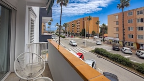 Un balcón con una silla blanca, vista a una calle con coches y palmeras, y edificios de varios pisos al fondo.
