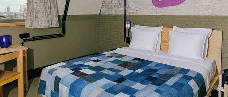 A bedroom with a checkered bedspread, a wooden nightstand, a desk with a lamp, and a city view through a window.