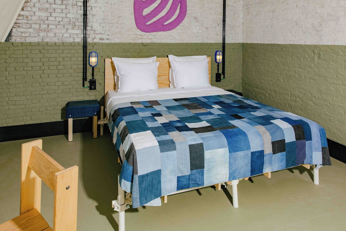 Una camera da letto con un letto in legno, una coperta a quadri blu e bianchi, due comodini e una parete con un motivo astratto viola.