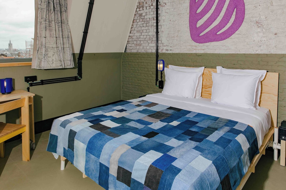 A bedroom with a checkered bedspread, a wooden nightstand, a desk with a lamp, and a city view through a window.