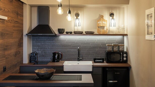 Een moderne keuken met een zwart metselwerk achterwand, een roestvrijstalen spoelbak, een magnetron en een koffieapparaat. Er zijn hangende lampen en een houten bord boven de spoelbak.