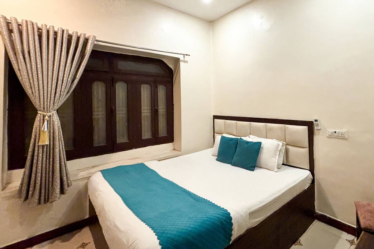 A neatly made bed with a blue accent, a window with curtains, a ceiling fan, and a wall socket.