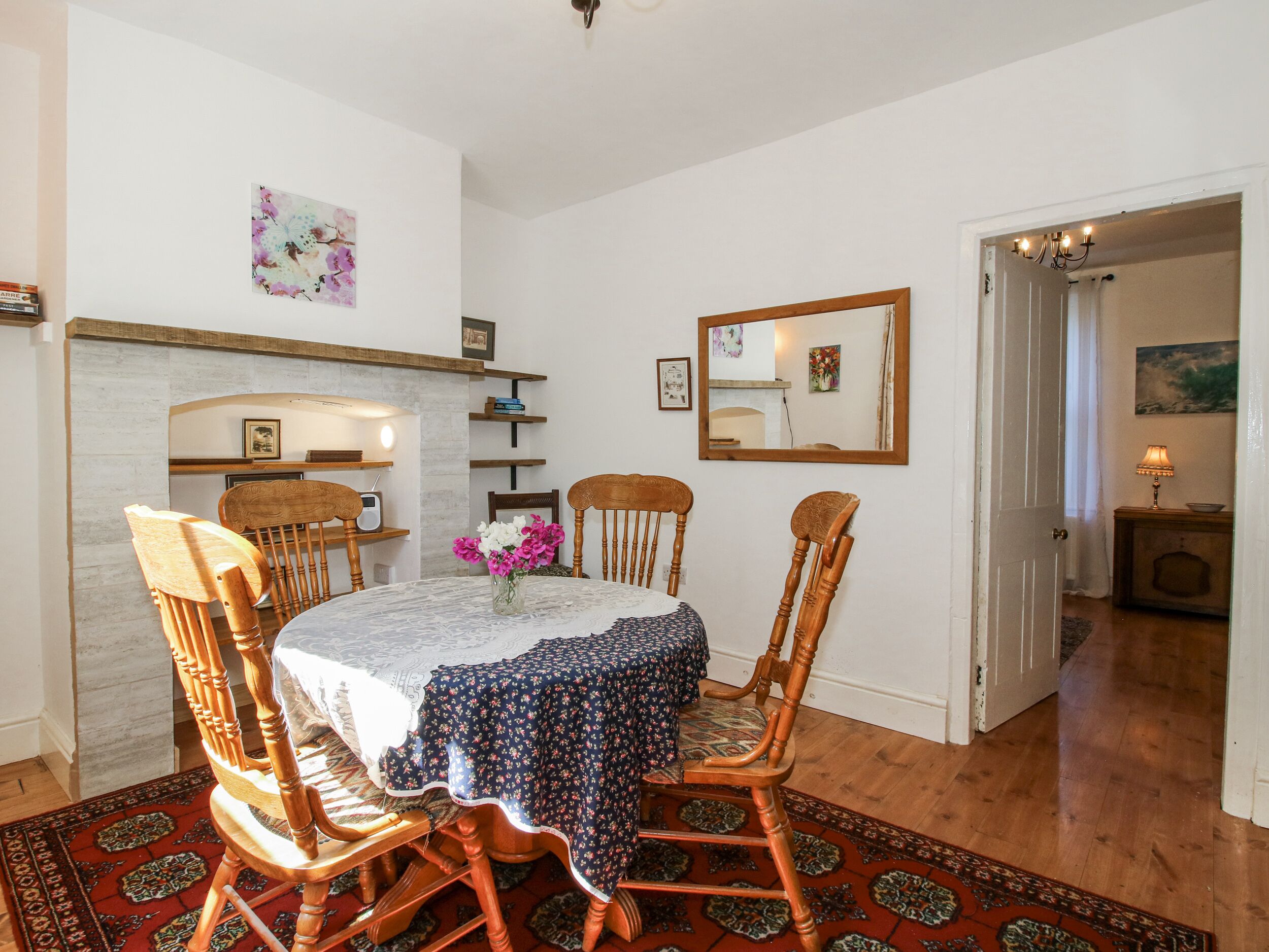 A dining area with a round table, four wooden chairs, a floral tablecloth, a vase with flowers, and a mirror on the wall.