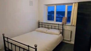 Une petite chambre avec un lit simple, une fenêtre, un radiateur et une armoire noire.