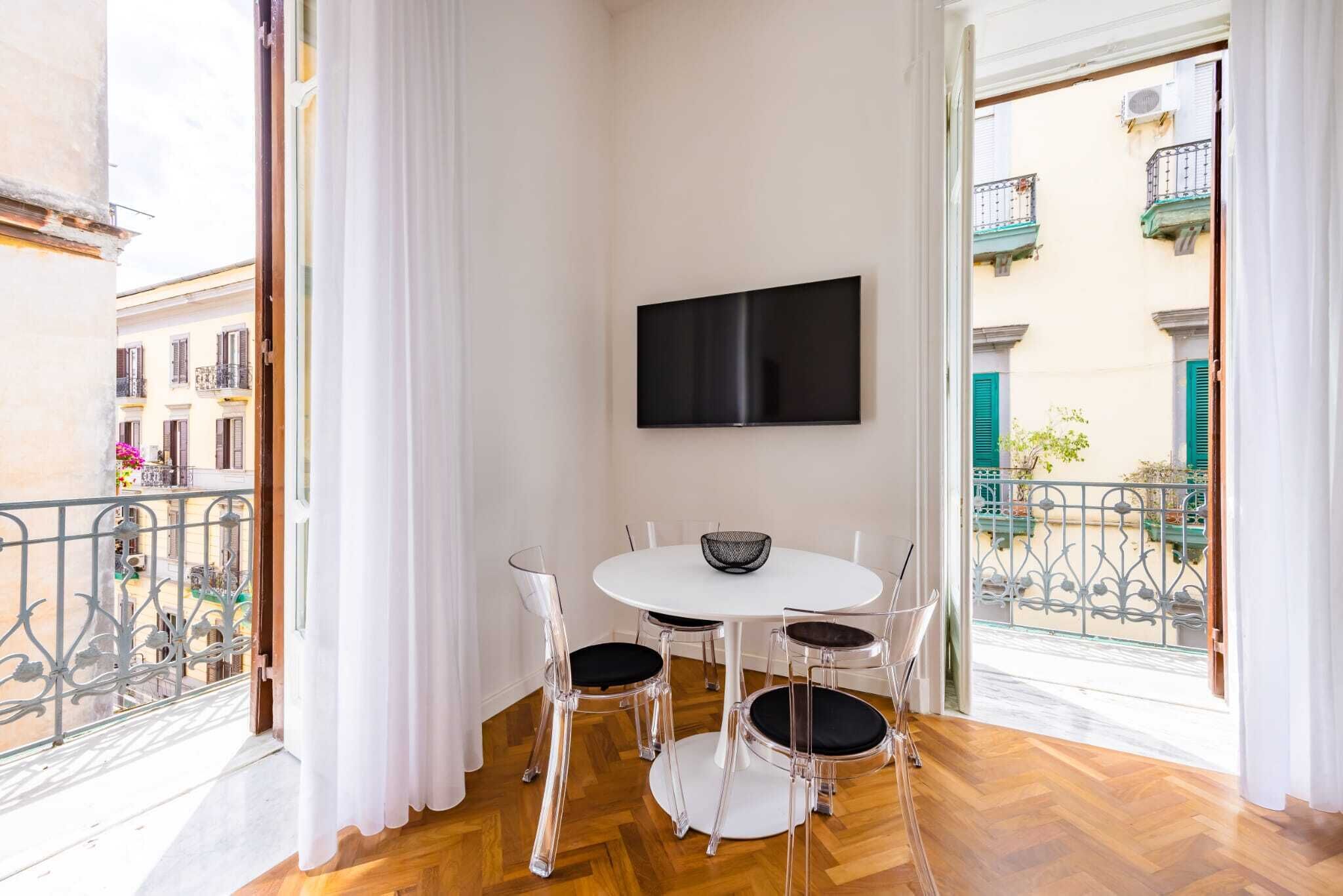 A dining area with a round table and four chairs, a flat-screen TV mounted on the wall, and a balcony with metal railings.