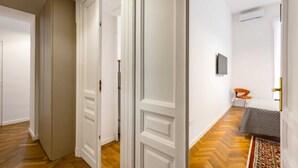 Un pasillo con puertas blancas, suelo de madera, una cama con colcha gris y una silla.
