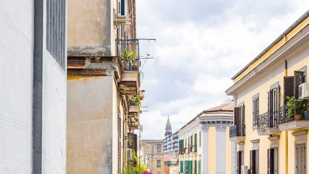 Una calle estrecha flanqueada por edificios, con balcones adornados con plantas y un cielo despejado.