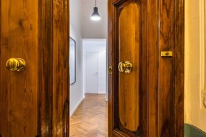 Une porte en bois, dotée de poignées en laiton, donne sur un couloir au sol en parquet à motifs en chevrons et où est suspendu un luminaire.