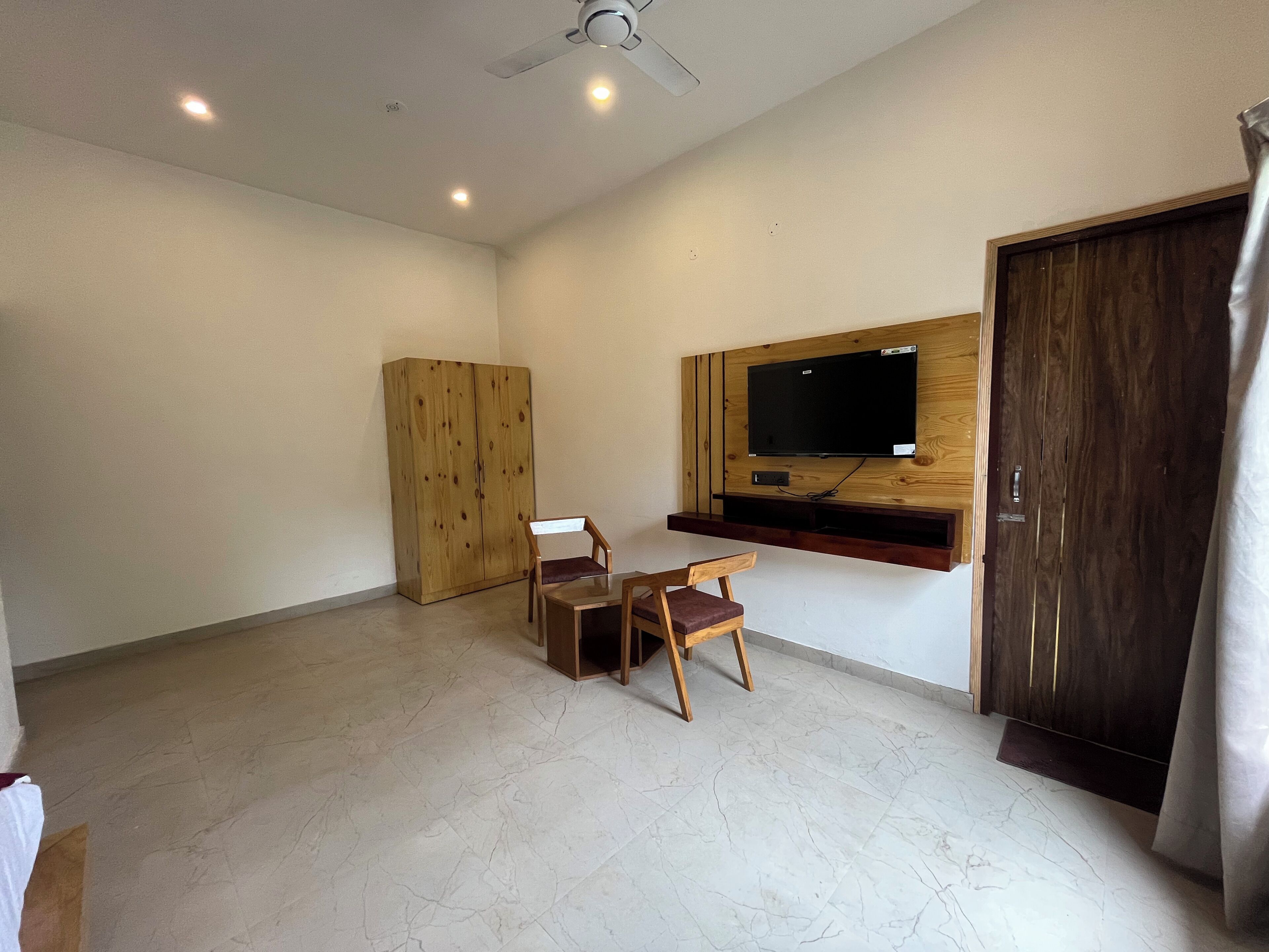 A room with a wooden wardrobe, a TV mounted on a wooden panel, a ceiling fan, and a tiled floor.