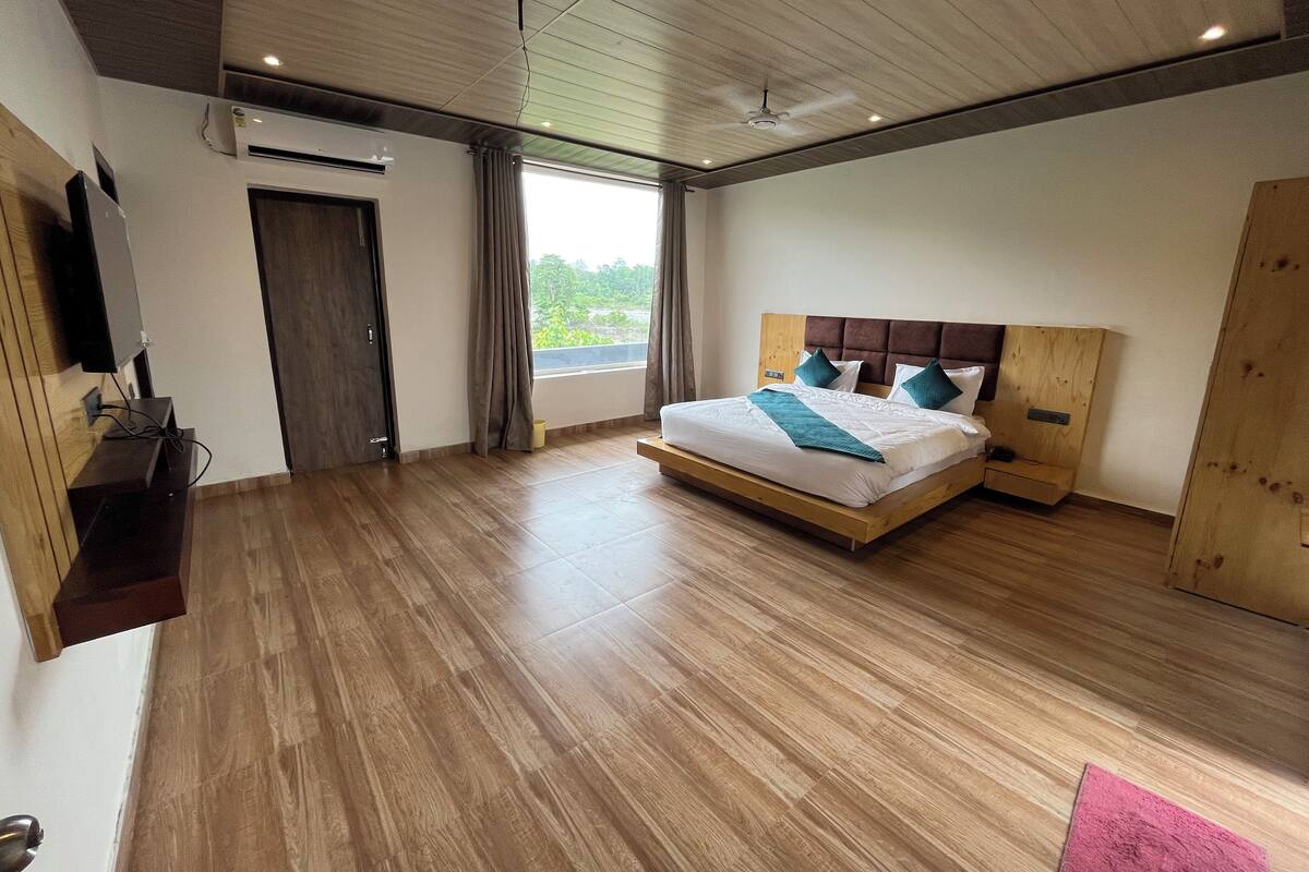 Ein modernes Schlafzimmer mit Holzboden, einem Bett mit Kopfteil, einem an der Wand montierten Fernseher und einem Fenster mit Vorhängen.