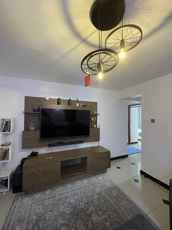 Un salon moderne doté d’un grand téléviseur à écran plat fixé sur un meuble mural en bois, d’un tapis à motifs et d’un luminaire au plafond original.
