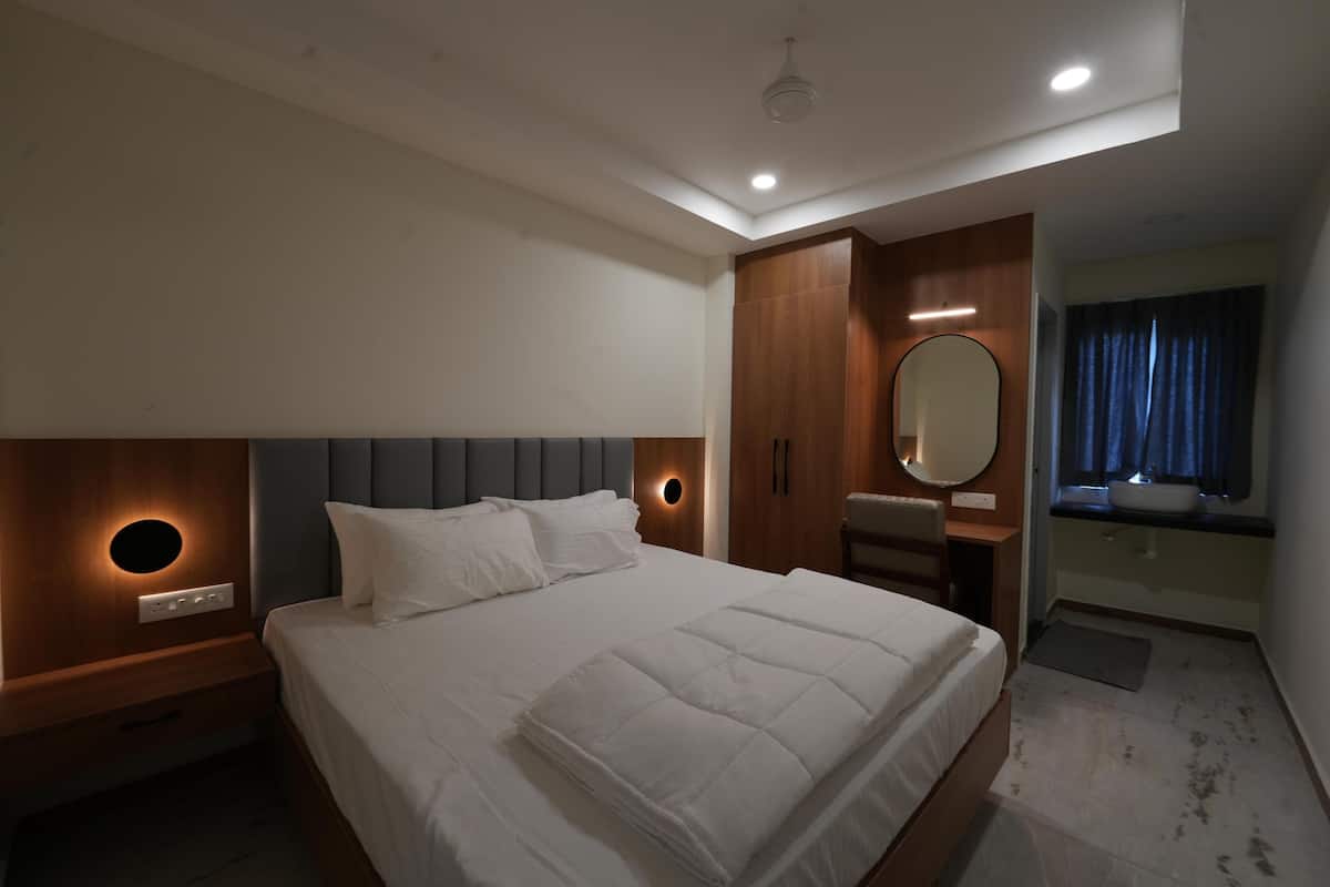 A modern hotel room with a large bed, a bedside table, a built-in wardrobe, a round mirror, and a bathroom with a sink and window.