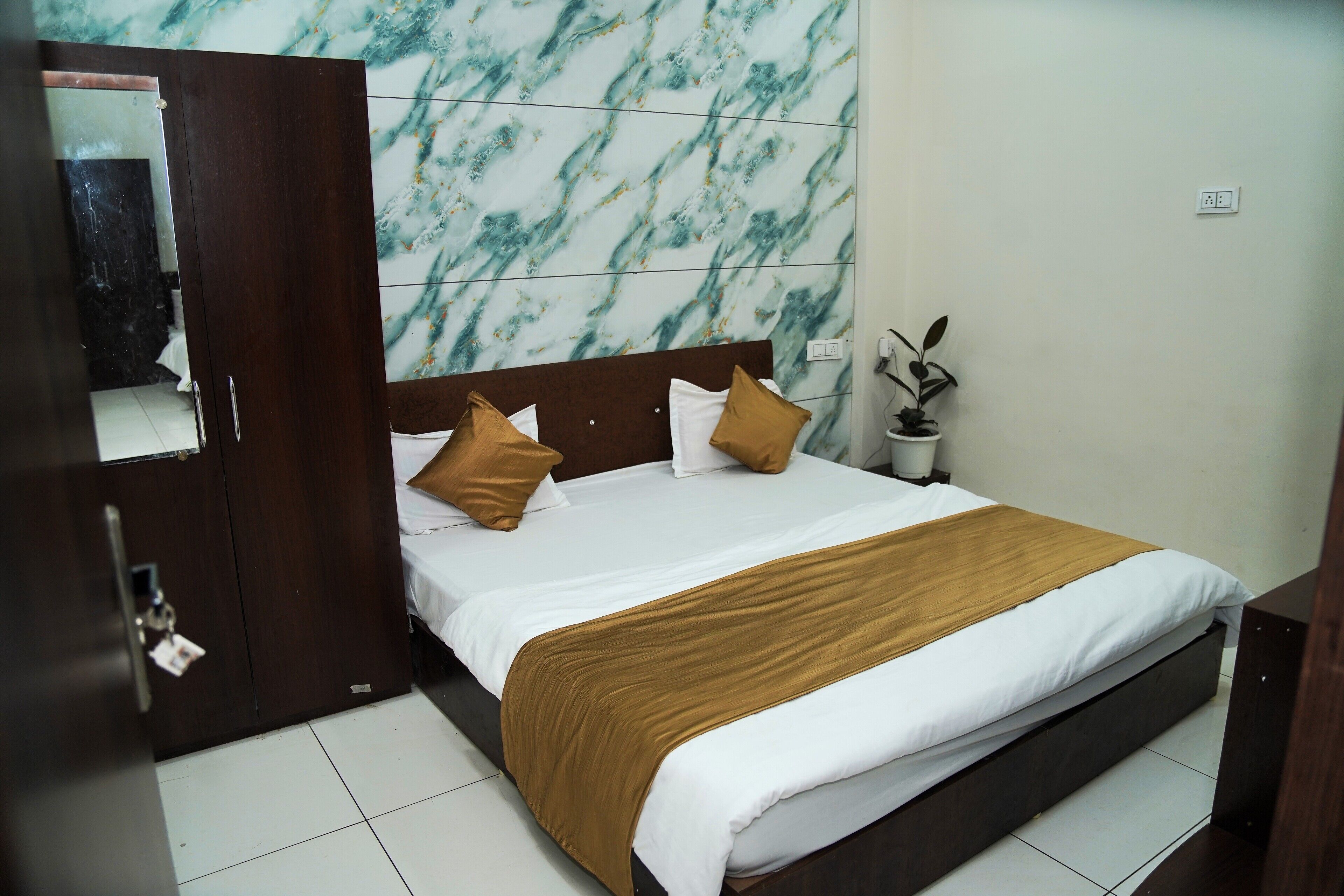 A hotel room with a bed, a wooden wardrobe, a marble-patterned wall, and a potted plant.