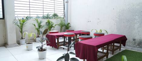 A room with tiled flooring, potted plants, tables with red tablecloths, and a window with blinds.