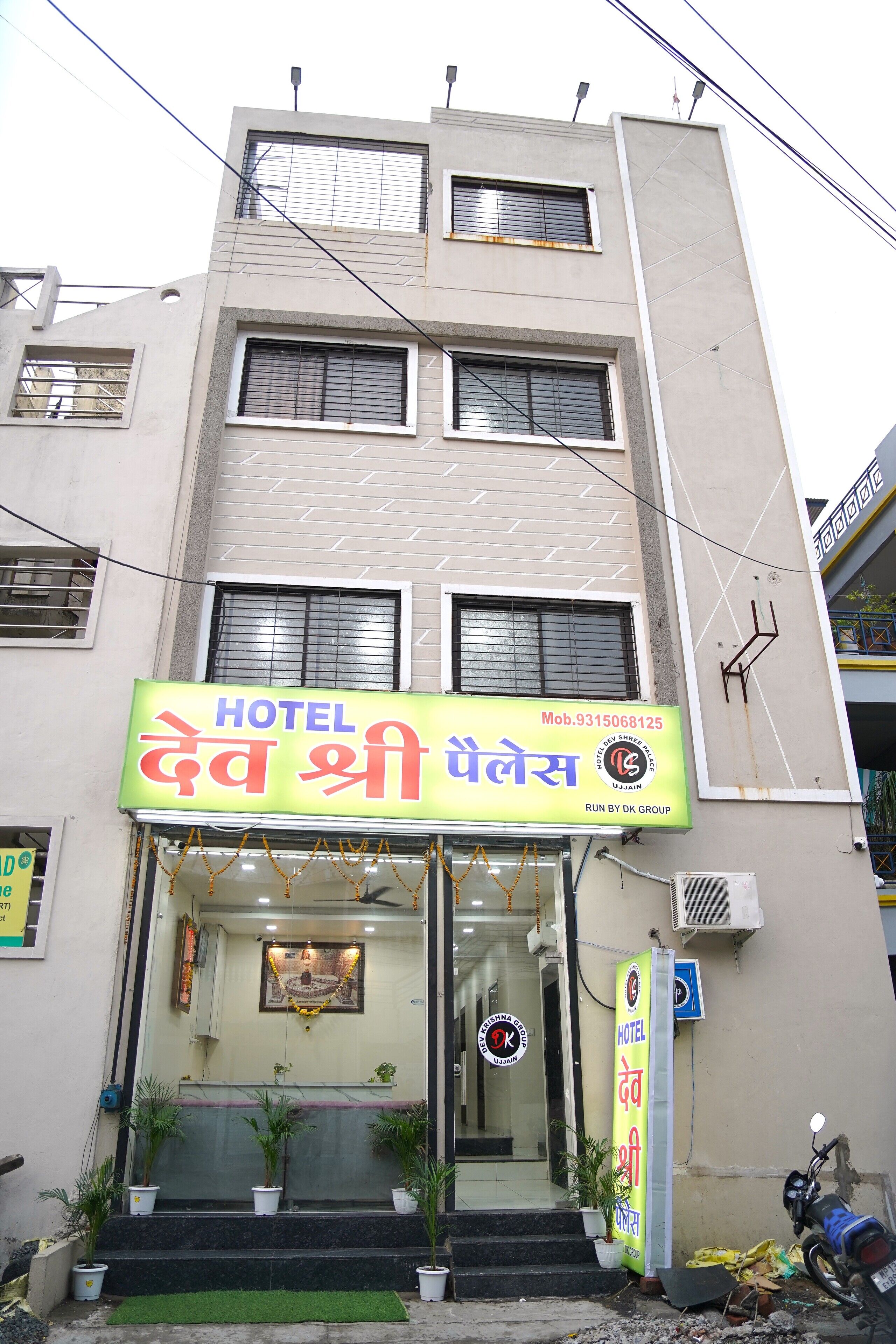 Ein Hotel namens "Hotel Dev Shri Palesh" mit Schild, Eingang und Blumentöpfen davor.