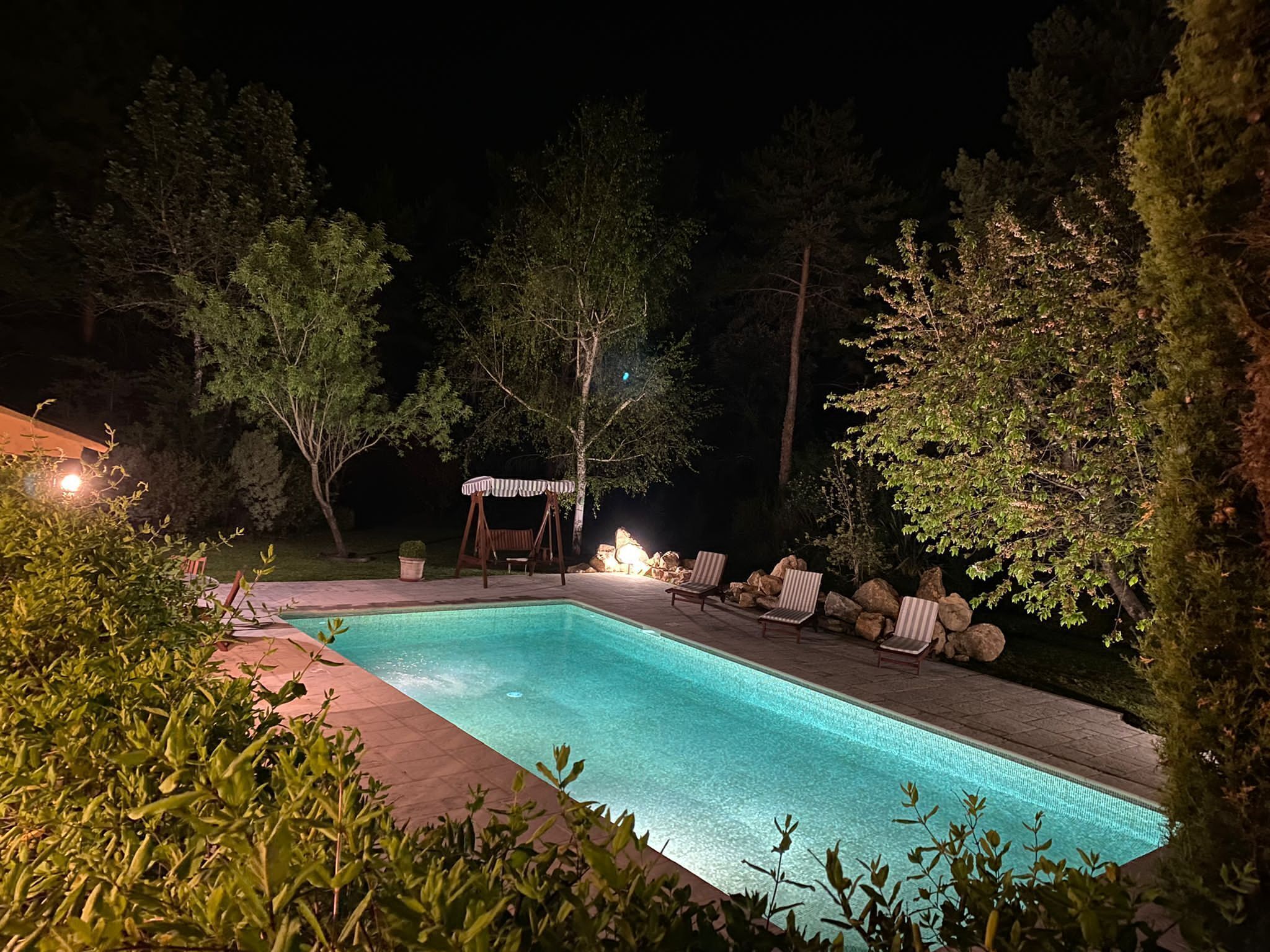 Piscina rectangular con solárium empinado y baldosas, rodeada de árboles y mobiliario de exterior.