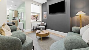 Een moderne woonkamer met een ronde salontafel, grijze banken, een flatscreen televisie aan de muur en een lamp op een bijzettafeltje.