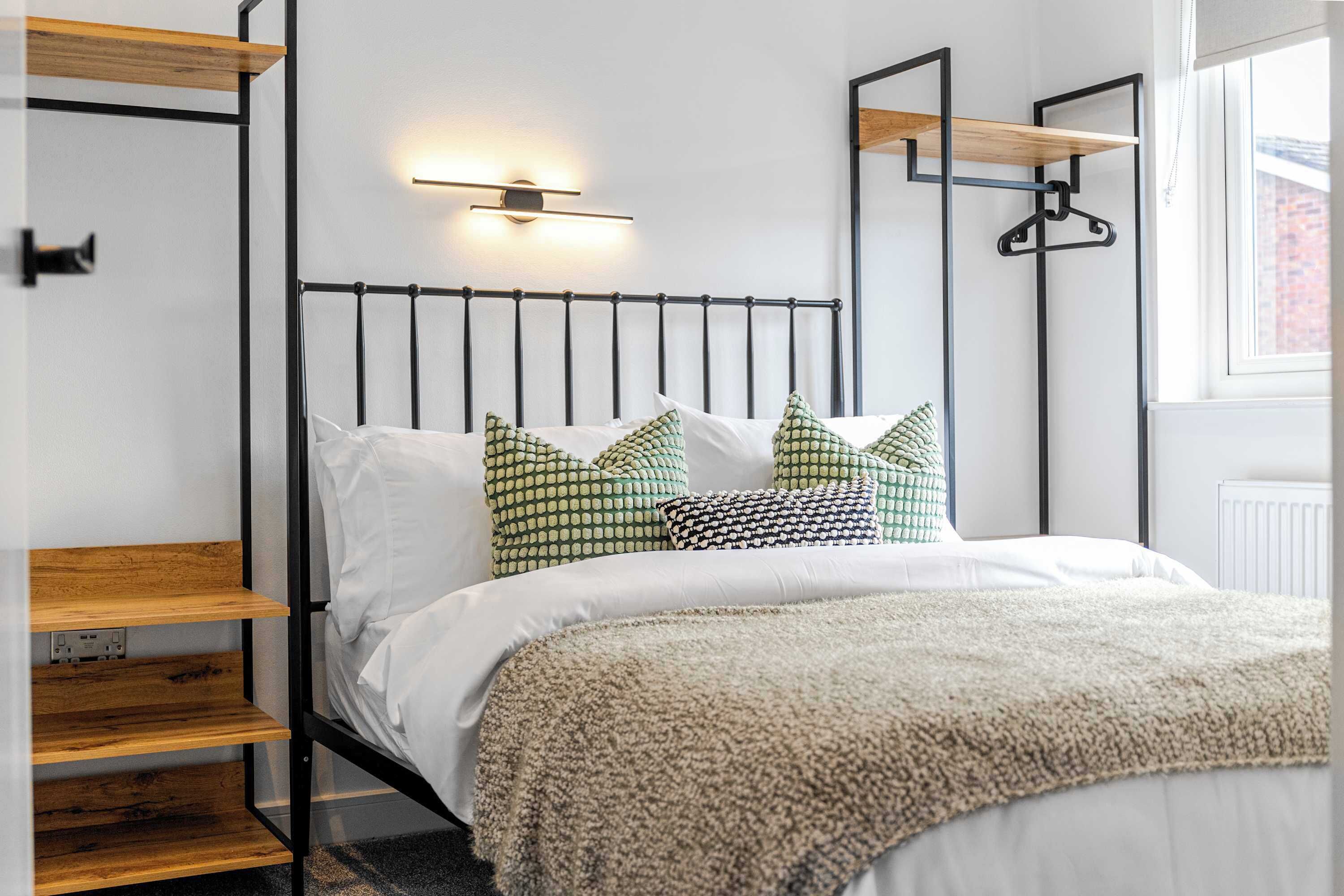 Un lit avec un cadre en métal, une literie blanche et des coussins à motifs. Il y a un luminaire fixé au mur et une étagère en bois.
